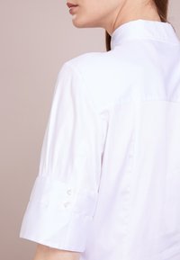 Camicia bianca in cotone con colletto alla coreana, maniche corte e due dettagli con bottone su ciascun polsino, caratterizzata da una texture liscia e una vestibilità sartoriale.