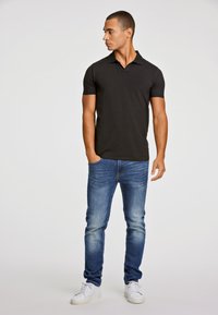 Svart kortärmad polo skjorta, figursydd design. Buren med ljusblå slim-fit jeans och vita sneakers, stående mot en neutral bakgrund.