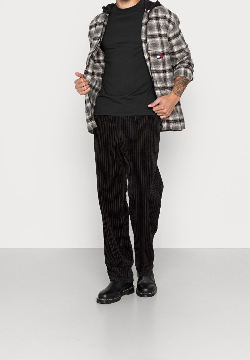 Zwarte corduroy broek met verticale strepen, gecombineerd met een grijs geruit shirt en een zwart t-shirt. De model draagt zwarte leren laarzen.