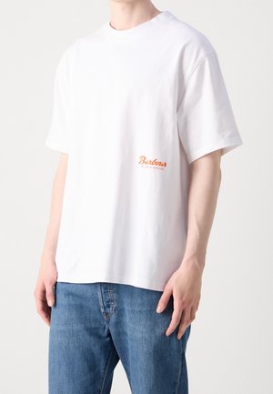 Osoba ubrana w gładki biały t-shirt z krótkim rękawem i pomarańczowym logo „Barbour of South Shields” na lewej piersi, w zestawieniu z niebieskimi dżinsami.