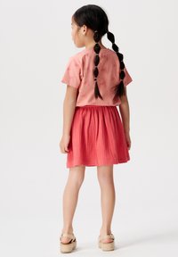 Roze shirt met korte mouwen, bedekt met een herhalend tekstpatroon, gecombineerd met een knielang rode textuurrok en neutrale espadrille sandalen.