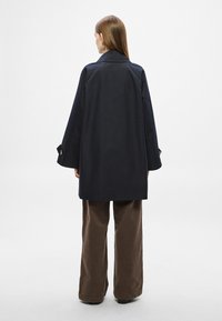 Παλτό σε ναυτικό μπλε, υπερμεγέθες με ευρύ γιακά, μακριά μανίκια και διακοσμητικά κουμπιά, συνδυασμένο με καφέ παντελόνι φαρδιάς γραμμής. Λεία υφή, μινιμαλιστικός σχεδιασμός.