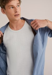 Tanktop in lichtgrijs met een gestructureerd patroon, gelaagd onder een zachtblauwe blouse met knoopjes. De blouse heeft opgerolde mouwen en een ontspannen pasvorm.