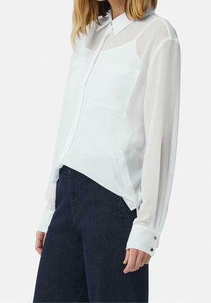 Personne portant un chemisier blanc à manches longues avec deux poches poitrine et un pantalon taille haute foncé sur un fond uni.