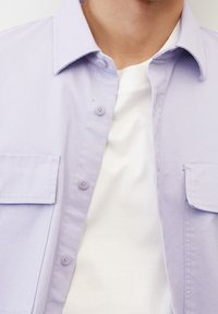 Camicia button-up di un leggero colore viola con due tasche frontali, colletto e maniche corte, indossata sopra una maglietta bianca a girocollo. Texture liscia.