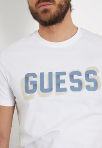 Balta medvilninė marškinėliai su dideliu mėlynu užrašu "GUESS" ir šviesiu smėlio spalvos šešėliu, apykaklė su apvaliu iškirpimu, trumpomis rankovėmis, vientisa medžiaga.