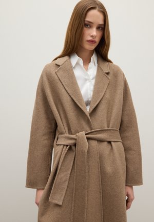 Abrigo largo con cinturón en lana de color marrón claro, con un cuello con muescas, mangas amplias y una textura suave. Estilizado sobre una camisa blanca.