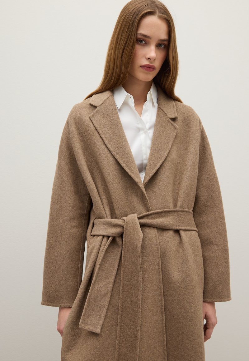 Cappotto lungo a cintura in lana marrone chiaro, con colletto a risvolto, maniche ampie e una texture morbida. Indossato sopra una camicia bianca.