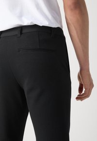 Homme portant un pantalon habillé noir et une chemise blanche, montrant le dos et le côté avec un gros plan sur la poche et le bras.