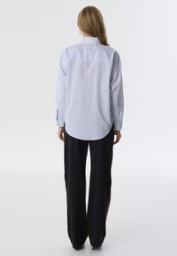 Camicia a righe blu e bianca, vista posteriore, con maniche lunghe e orlo arrotondato. Abbinata a pantaloni neri con dettagli in bronzo.