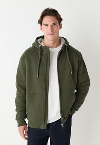 Olijfgroene hoodie met rits, gevoerd met fleece aan de binnenkant, capuchon met trekkoord, zakken aan de voorkant en geribde manchetten, gedragen over een wit shirt.