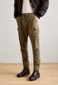 Olivgrüne Cargo-Hosen mit elastischem Bund und Kordelzug, ausgestattet mit Seitentaschen und schmal zulaufenden Bündchen. Getragen mit braunen Stiefeletten.