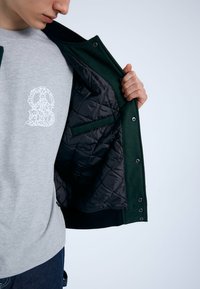 Giacca verde con una vestibilità trapuntata nera e bottoni a pressione, indossata sopra una camicia grigia chiara con un motivo floreale bianco.