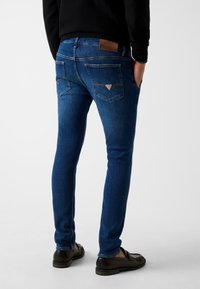 Dopasowane ciemnoniebieskie jeansy z gładką teksturą, pięcioma kieszeniami i skórzaną naszywką na pasku do spodni. Akcenty w postaci kontrastującego przeszycia.