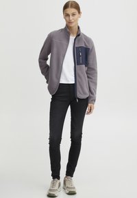 Grijze fleece jack met een marineblauwe borstzak, met rits. Draag het over een wit shirt, gecombineerd met zwarte jeans en grijze sneakers.