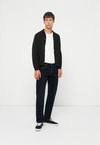 Ung mann som står, iført svart knappet cardigan, hvit t-skjorte, mørke jeans og svart-hvite sneakers mot hvit bakgrunn.