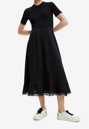 Schwarzes Kleid mit einem gerippten, kurzärmeligen Oberteil und einem strukturierten Rock mit einer Spitzenkante am Saum, kombiniert mit weißen Sneakers.