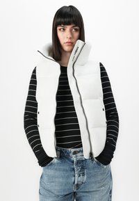 Vrouw met steil donker haar, gekleed in een witte puffer vest, een zwart-wit gestreepte lange mouwen top en blauwe jeans, met haar handen in de zakken.
