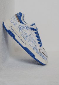 Zapatilla blanca con acentos azules, que presenta ilustraciones detalladas de paisajes en líneas azules y texto manuscrito en el lateral.