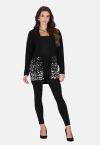 Schwarze Strickjacke mit Paillettenverzierung am Saum, offenes Design, getragen über einem enganliegenden schwarzen Oberteil und Leggings, kombiniert mit schwarzen Schuhen.
