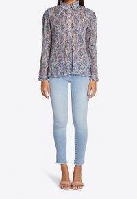 Blusa floral de manga larga en tonos azul, rosa y verde; con cuello, abotonada y puños con volantes; llevada con jeans ajustados de un azul claro.
