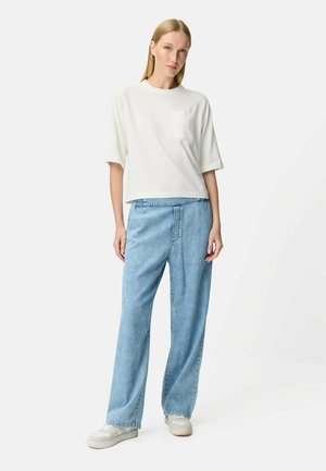 Femme blonde debout portant un haut blanc à manches courtes avec poche, un jean large bleu clair et des baskets blanches sur un fond blanc.