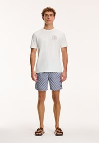 Balta trumpa rankovė t-shirt su apvaliu iškirpimu ir mažuoju logotipu ant krūtinės, derinama su mėlynomis raštuotomis maudymosi šortais ir riešutų spalvos šlepetėmis.