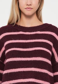 Pull en tricot rayé bordeaux et rose avec un col large côtelé et une coupe ample. Comprend un matériau en mélange de laine douce et texturée.