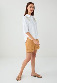 Chemise blanche à manches courtes avec broderie florale dorée et col, assortie à un short marron clair et des tongs.