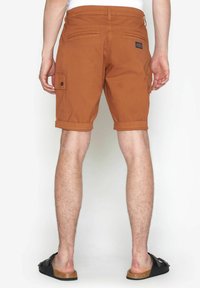 Pantaloni scurți cargo din bumbac maro, cu margini rulante, buzunare laterale și buzunare din spate cu patch. Textură netedă și design casual.