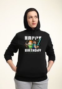 Felpa nera con cappuccio caratterizzata da un grande grafico con personaggi pixelati e il testo "BUON COMPLEANNO". Tessuto morbido, tasca a marsupio, cordino regolabile.