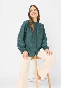Teal, textured button-up shirt with long sleeves, featuring wavy patterns. Paired with light-colored pants, model seated on a wooden stool.