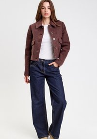 Braune kurze Jacke aus strukturiertem Stoff, mit Kragen, silbernen Knöpfen und zwei Vordertaschen, kombiniert mit weitgeschnittenen Jeans aus dunklem Denim.