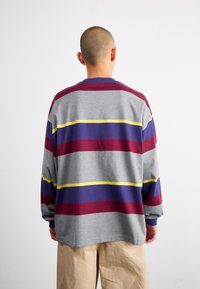 Pelēks garām piedurknēm krekls ar izteiksmīgām bordo, purpura un dzeltenām horizontālajām strīpām. Brīvā piegriezumā ar apaļu izgriezumu un ar kājām aizsietām piedurknēm.