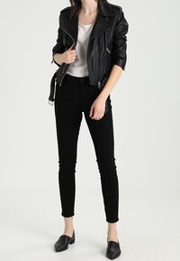 Schwarze Lederbikerjacke mit Reißverschlussdetails, kombiniert mit einem figurbetonten wei ßen Oberteil, schwarzen Skinny-Jeans und schwarzen Mokassins.