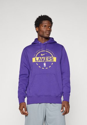 Mand iført en lilla Los Angeles Lakers hoodie med gult og hvidt logo og lysegrå bukser, stående mod en ensfarvet baggrund.