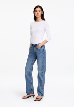 Femme debout les mains dans les poches, portant un haut blanc à manches longues, un jean bleu et des sandales marron à talons, sur fond blanc.