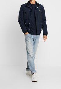 Marineblaues Jacket mit schwarzem Kragen, Reißverschluss und Druckknöpfen, mit zwei Fronttaschen. Getragen über einem marineblauen Hemd mit hellblauen Jeans.