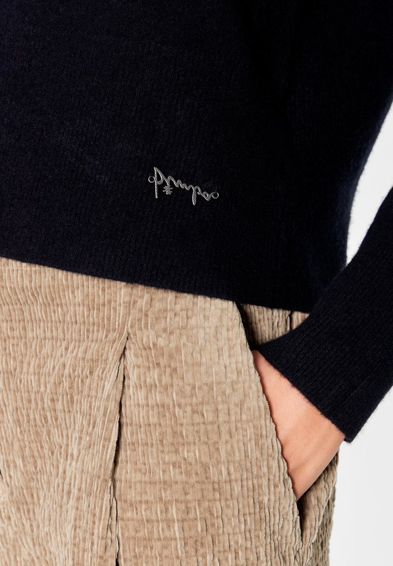 Ciemnogranatowy sweter wykonany z miękkiego materiału, z eleganckim srebrnym akcentem logo. W połączeniu z fakturowanymi beżowymi spodniami z korduru, które mają lekko pognieciony wygląd.