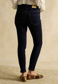 Personne portant un jean skinny bleu marine et des chaussures plates marron à motifs, debout devant un fond beige uni.