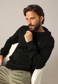 Haut à capuche noir texturé avec un motif matelassé, capuche à cordon de serrage et poignets côtelés, associé à un pantalon léger en kaki.