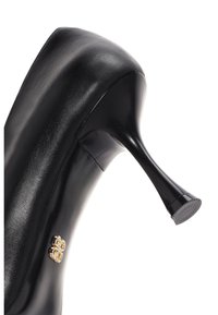 Zapato de tacón alto de cuero negro con un acabado brillante, punta afilada y tacón de aguja cónico. Presenta un adorno decorativo en tono dorado.