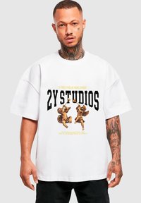 Fehér, oversize T-shirt, amelyen két cherub grafikája, vastag fekete felirat "2Y STUIDOS" és alatta sárga kiemelő szöveg található.