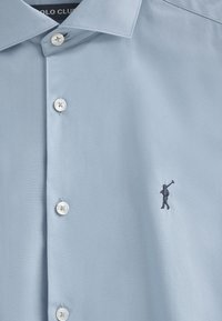 Lichtblauw overhemd met een zachte textuur, voorzien van een kleine geborduurde figuur die een golfclub vasthoudt en vier witte knopen langs de voorkant.