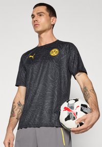 Sort mønsteret atletisk skjorte med korte ærmer og gul logo. Modellen holder en sort og hvid fodbold med rød Puma-logo. Grå shorts.