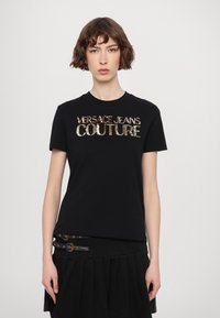 Črna bombažna majica s kratkimi rokavi, ki vsebuje zlato bleščičasto logo "VERSACE JEANS COUTURE". Prilegajoča zasnova s klasifikacijo določenega okroglega izreza.