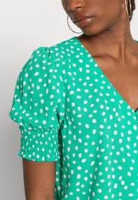 Blusa verde com bolinhas brancas, apresentando um decote em V profundo, mangas bufantes e detalhes de punhos franzidos. Tecido liso e leve.