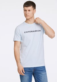Vaaleansininen puuvillainen T-paita, jossa on painettu "COPENHAGEN" -logo tummalla tekstillä. Pyöreä kaula-aukko, lyhyet hihat, yhdistettynä farkkuhousuihin.