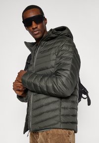 Muškarac u tamnozelenoj jakni s kvadratićima, s crnim sunčanim naočalama i smeškim hlačama od kordura, prilagođava remen crnog ruksaka na bijeloj pozadini.