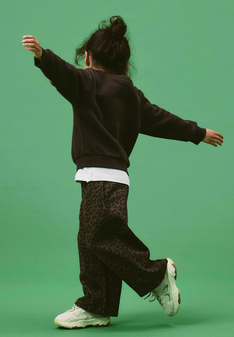 Enfant aux cheveux foncés en chignon, portant un sweat-shirt noir, une chemise blanche, un pantalon imprimé léopard et des baskets blanches, en équilibre sur un pied.